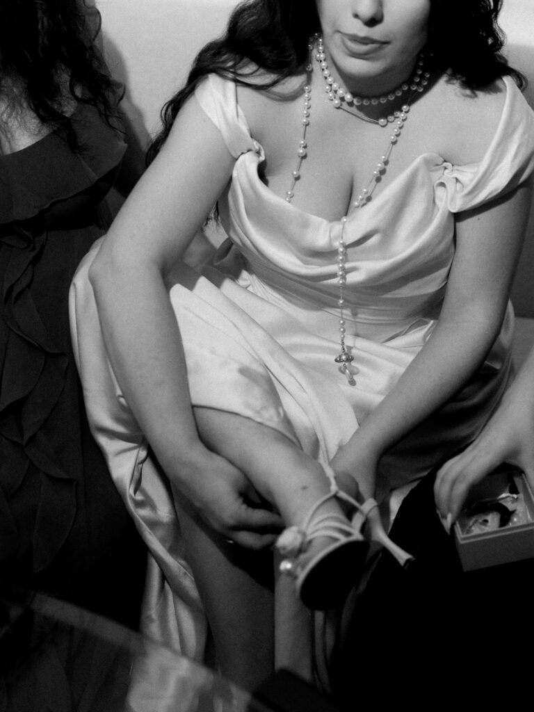 A black-and-white close-up of the bride in her white satin cowl-neck wedding gown being helped with her gloves, a long pearl and rosary cross necklace draped elegantly across her décolletage, captured in an intimate getting-ready moment at One&Only The Palm Dubai wedding.