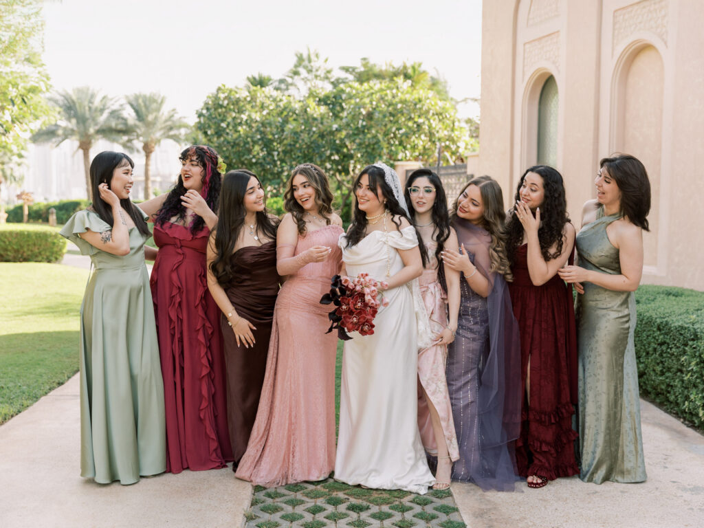 The bride in her white wedding gown and cathedral veil stands surrounded by seven bridesmaids in mismatched floor-length gowns in rich jewel tones — burgundy, sage, blush, mauve, and dusty blue — laughing and looking at her adoringly in the lush garden grounds of One&Only The Palm Dubai, with the resort's arched cream stone facade and tropical greenery behind them.