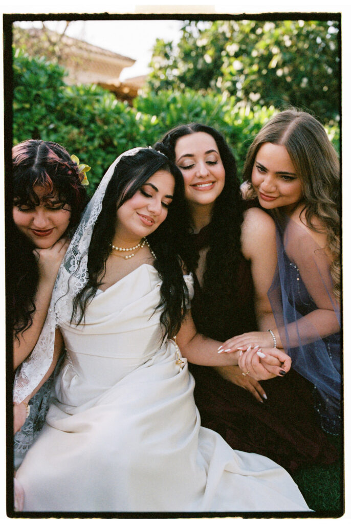 A film photograph of the bride in her white cowl-neck wedding gown and lace cathedral veil sitting closely with three bridesmaids in jewel-toned gowns, all leaning in together with eyes closed in a warm, tender bridesmaid moment in the gardens of One&Only The Palm Dubai.