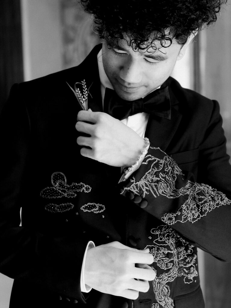A black-and-white close-up detail shot of the groom's custom black tuxedo jacket, showcasing intricate hand-embroidered cloud and floral motifs across the lapel and sleeve, with a wheat boutonniere pinned to the chest — a nod to his identity as a Disney animator.