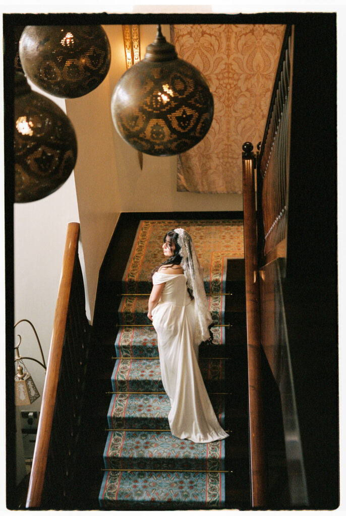 A warm film photograph of the bride in her white satin off-the-shoulder wedding gown and lace cathedral veil pausing midway on an ornate tiled staircase inside One&Only The Palm Dubai, looking back over her shoulder, framed dramatically by two large hanging Moroccan brass lanterns and richly patterned wallpaper above her.
