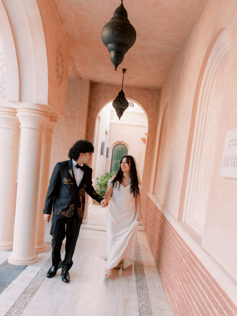 The bride and groom walk hand in hand through a stunning rose-pink arched colonnade corridor at One&Only The Palm Dubai, a large ornate Moroccan brass lantern hanging above them, the bride's white satin gown trailing behind her as they look at each other tenderly, with the resort's signature blush archways receding elegantly into the background.