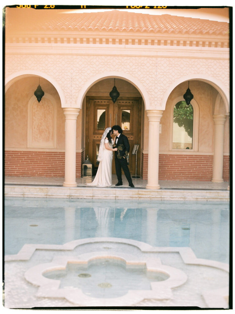 A film photograph of the bride and groom sharing a romantic embrace beside a geometric Moorish reflecting pool at One&Only The Palm Dubai, the still water mirroring the resort's warm terracotta arched facade and ornate wooden doors behind them in the soft afternoon light.
