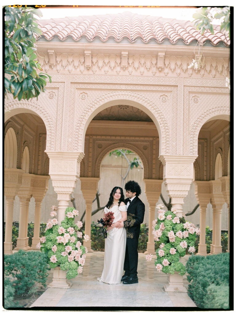 The bride and groom stand together beneath the ornate Moorish arched colonnade of One&Only The Palm Dubai, framed by a grand blush floral arrangement of garden roses and greenery, the bride in her white gown and lace cathedral veil and the groom in his custom gold-embroidered black tuxedo, with the resort's intricately carved cream stone facade glowing warmly behind them.