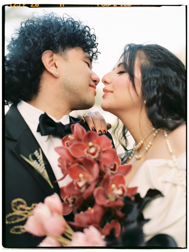 A dreamy close-up film portrait of the bride and groom leaning in for a kiss, noses almost touching, the bride's pearl necklace and deep red orchid bouquet filling the foreground with rich colour, the groom's curly hair and black bow tie visible as they share an intimate moment at the One&Only The Palm Dubai wedding.