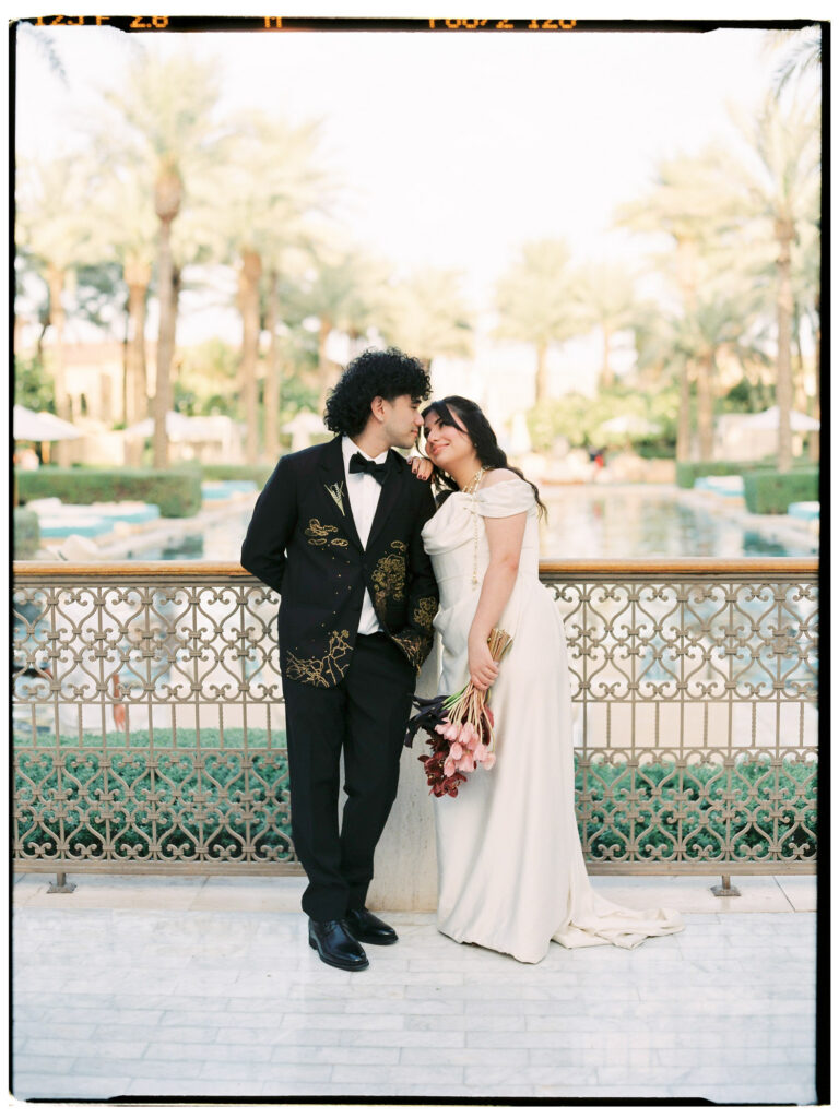 The bride and groom share a tender nose-to-nose moment on an ornate iron-railed terrace at One&Only The Palm Dubai, the bride in her white off-the-shoulder satin gown holding her deep red orchid bouquet, the groom in his custom gold-embroidered black tuxedo, with lush tropical palm trees and the resort's pool visible behind them.
