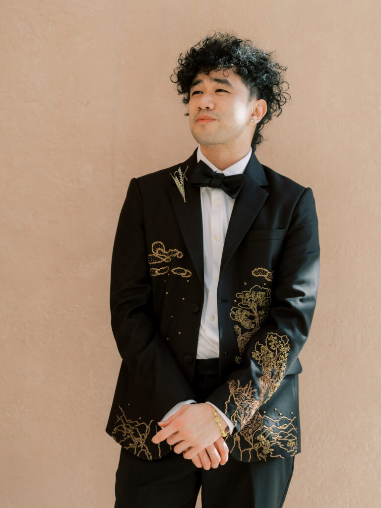 The groom stands confidently against a warm beige wall in his custom black tuxedo jacket featuring stunning gold embroidered cloud and botanical details along the lapel and cuffs, his curly hair and black bow tie completing the artistic, one-of-a-kind look at the One&Only The Palm Dubai wedding.