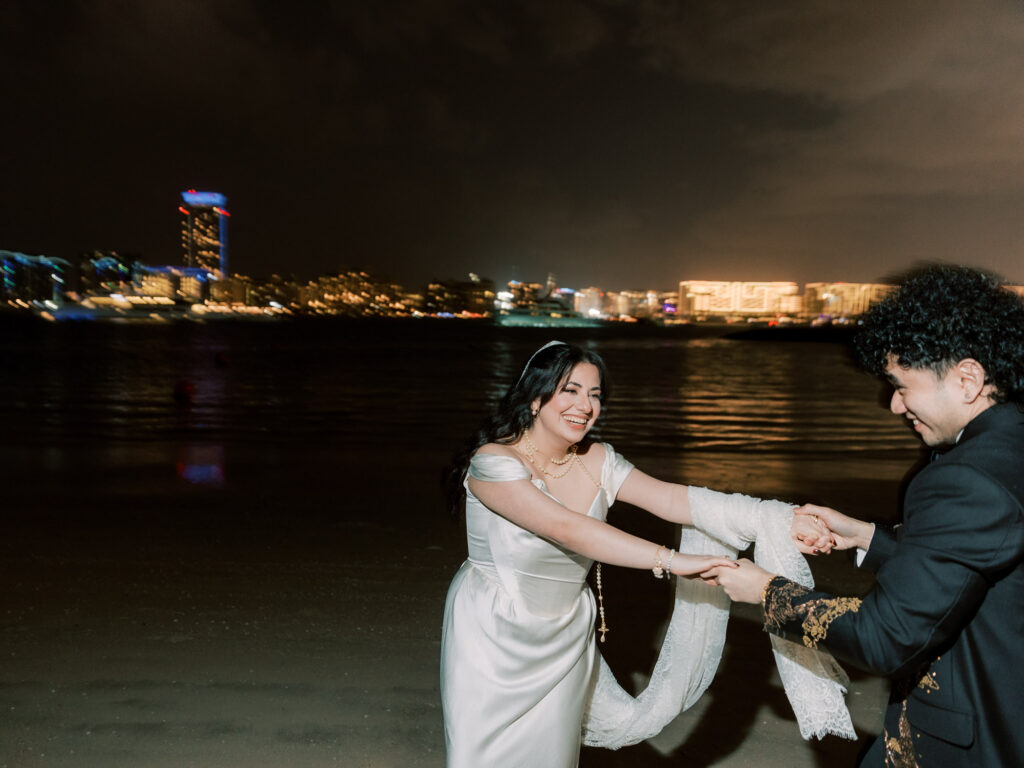 The bride and groom laugh and spin together holding both hands on the Dubai waterfront at night, the bride in her white off-the-shoulder satin gown and cathedral veil, the groom in his custom embroidered black tuxedo, with the colourful lit-up Dubai skyline stretching dramatically across the water behind them.