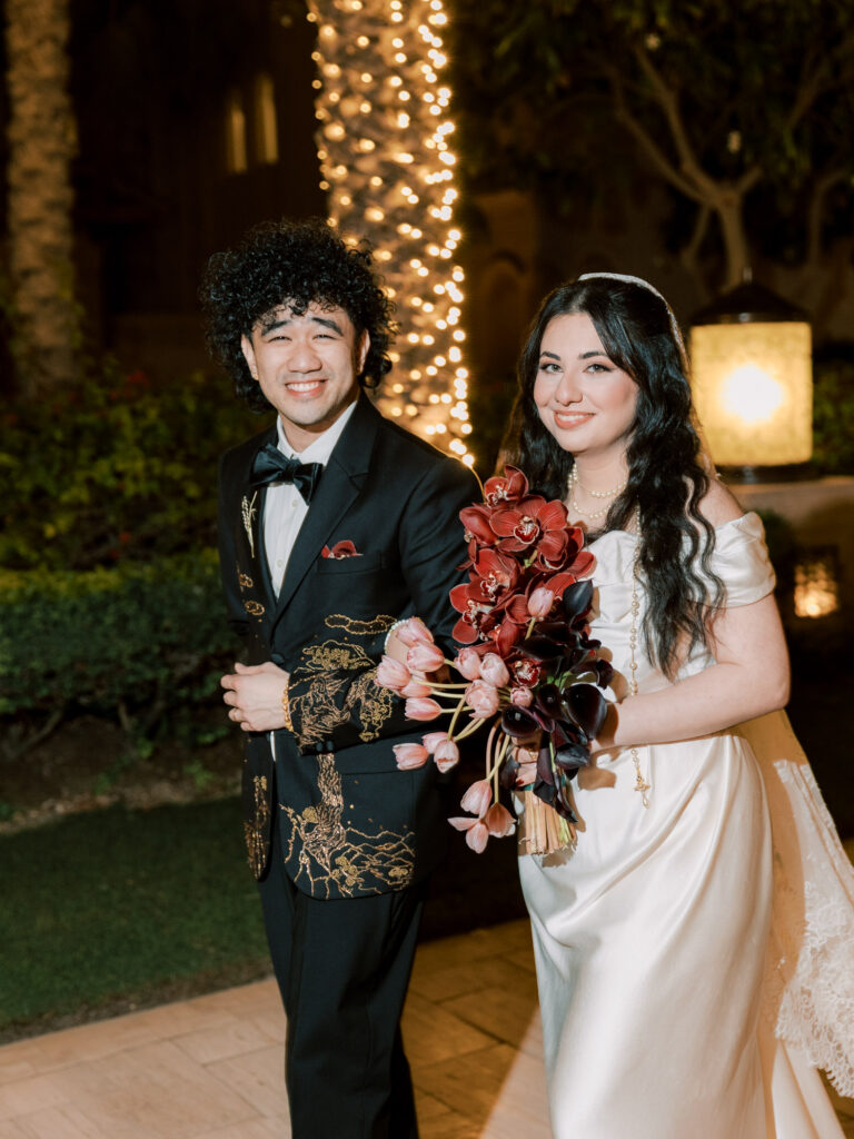 A joyful nighttime portrait of the bride and groom beaming at the camera in the illuminated gardens of One&Only The Palm Dubai. The groom's custom black tuxedo with intricate gold botanical embroidery is showcased beautifully alongside the bride in her white off-the-shoulder satin gown and pearl necklace, holding a lush bouquet of deep red cymbidium orchids, pink tulips, and black calla lilies, with fairy-light-wrapped trees and warm lanterns glowing softly behind them.