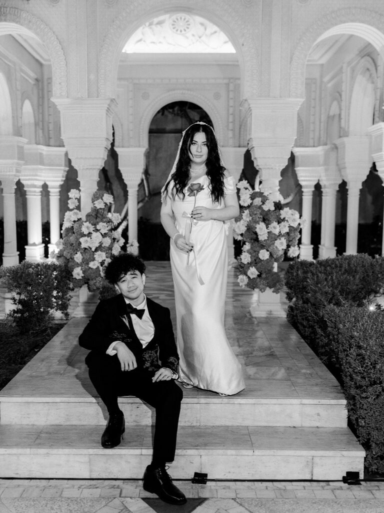 A black-and-white night photograph of the bride standing tall on the steps of One&Only The Palm Dubai's grand illuminated entrance pavilion, holding a single red rose, while the groom sits casually on the steps below her looking up, with large blush floral arrangements and the resort's ornate Moorish arched columns framing them dramatically.