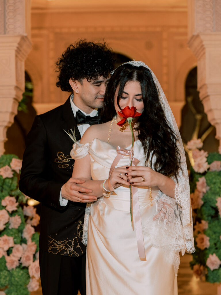 The groom stands behind the bride in the illuminated Moorish courtyard of One&Only The Palm Dubai at night, his arms wrapped around her as she holds a single red rose to her lips, both looking down tenderly, surrounded by blush floral arrangements and the resort's ornate cream stone arched facade glowing warmly around them