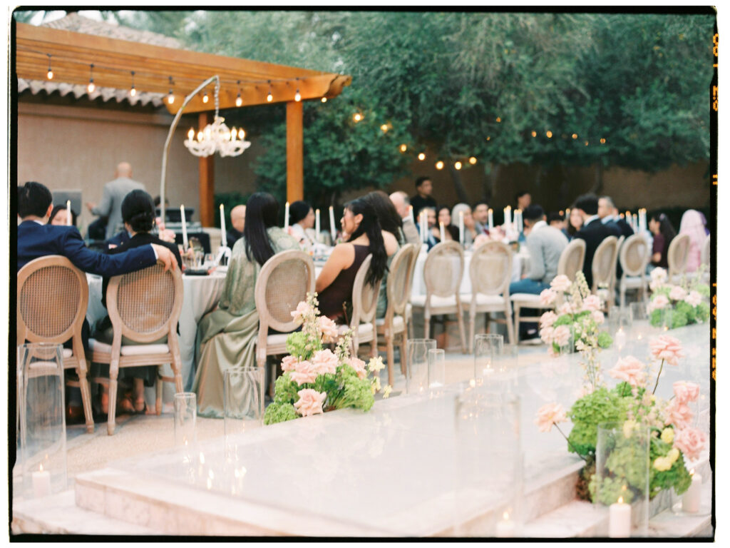 A wide film photograph of the full outdoor wedding reception at One&Only The Palm Dubai, with guests seated at long banquet tables in rattan Louis chairs beneath a wooden pergola hung with a crystal chandelier and string lights, surrounded by towering lush trees and blush floral centrepieces glowing in the warm evening light.