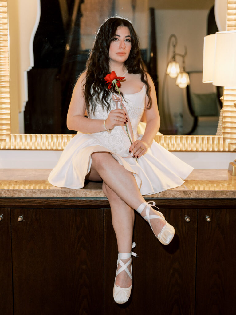 A Super 8mm-style photograph of the bride in a short white lace mini dress and white ballet flats sitting on a hotel dresser inside her One&Only The Palm Dubai suite, holding a single red rose and gazing directly at the camera with a cool, confident expression, warm lamp light glowing behind her.
