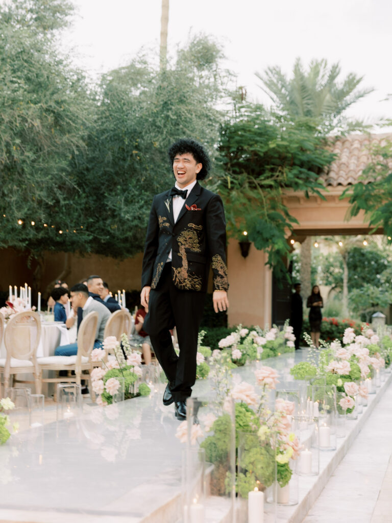 The groom beams with pure joy as he walks down the mirrored ceremony aisle at One&Only The Palm Dubai, wearing his custom gold-embroidered black tuxedo jacket and bow tie, laughing with uncontainable happiness as guests seated at round tables look on in the lush garden courtyard behind him.