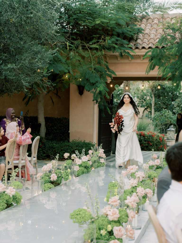 The bride walks down the reflective mirrored aisle toward waiting guests during the outdoor wedding ceremony at One&Only The Palm Dubai, her white satin gown and lace cathedral veil trailing elegantly behind her, the floral-lined aisle filled with blush roses, hydrangeas, and glass candle votives, with tropical trees and warm string lights glowing above.