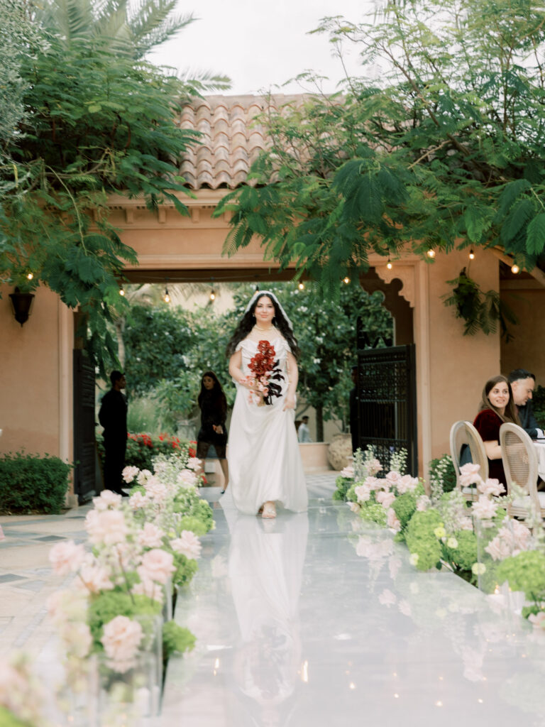 The bride in her sleek white cowl-neck satin wedding gown and lace cathedral veil walks down a reflective mirrored aisle lined with blush garden roses, greenery, and glass candleholders during her outdoor ceremony at One&Only The Palm Dubai, holding her deep pink and black bouquet, with lush overhanging trees, string lights, and the resort's terracotta-roofed pavilion framing the romantic scene.
