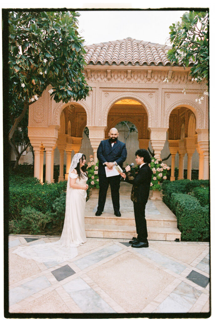 A film photograph of the bride and groom exchanging vows in the ornate Moorish-arched courtyard of One&Only The Palm Dubai, the groom reading from a microphone while the bride listens in her white off-the-shoulder satin gown and lace cathedral veil, with an officiant standing on the stone steps behind them, flanked by lush manicured hedges and large blush floral arrangements beneath the intricately carved cream stone facade.