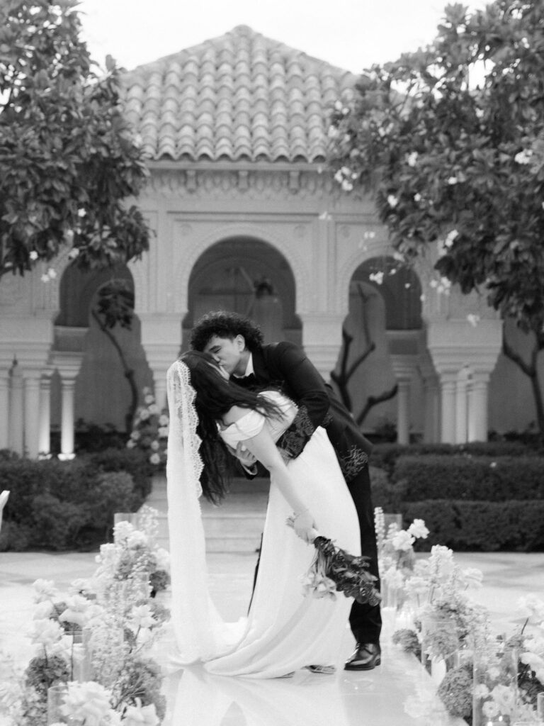 A dramatic black-and-white film photograph of the groom dipping the bride in a deep, sweeping kiss on the mirrored ceremony aisle at One&Only The Palm Dubai, her lace cathedral veil and satin gown trailing across the floor, surrounded by blush floral arrangements and the resort's ornate Moorish arched pavilion glowing behind them.