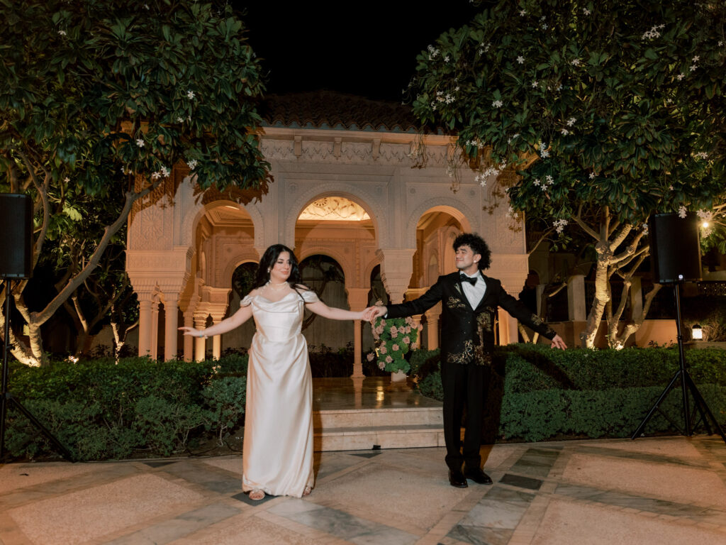 The bride and groom share their first dance in the illuminated outdoor courtyard of One&Only The Palm Dubai at night, spinning hand in hand beneath warm string lights and lush trees, the resort's ornate cream stone arched facade glowing beautifully behind them.