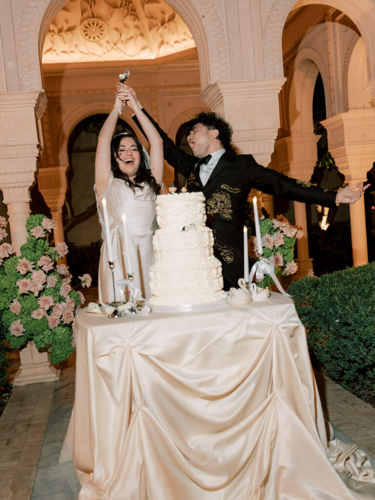 The bride and groom celebrate joyfully as they cut their elaborate white tiered wedding cake beneath the ornate Moorish arched pavilion of One&Only The Palm Dubai, both laughing and raising their arms in triumph, with blush floral arrangements and warm candlelight glowing around them.