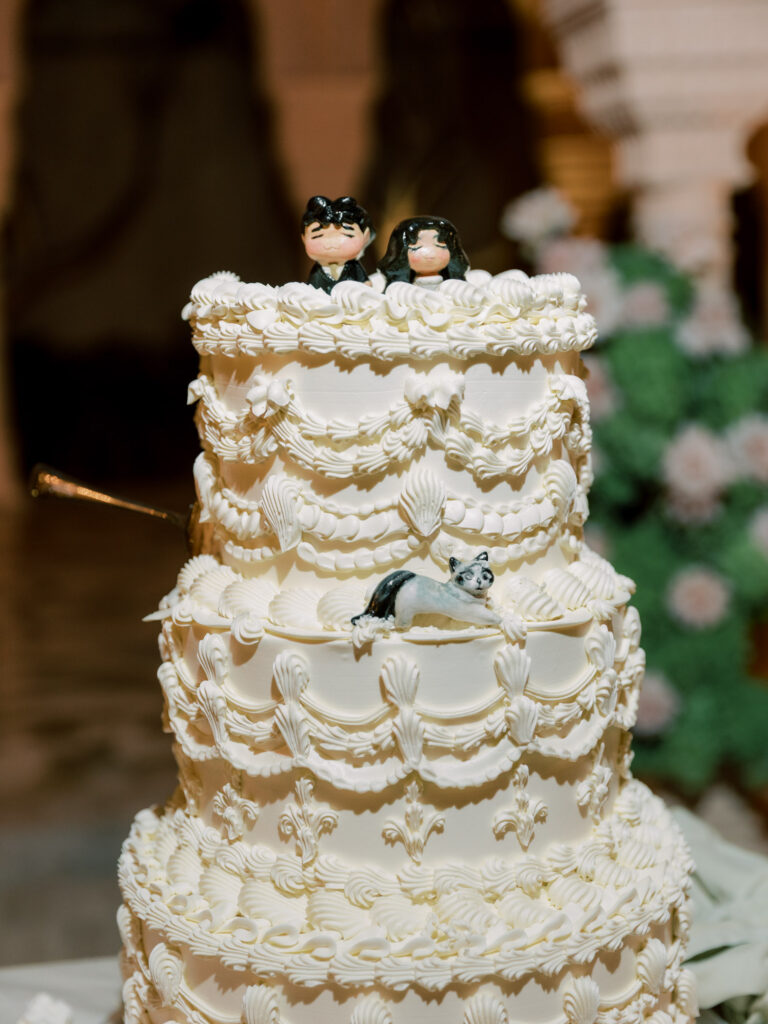A close-up detail shot of the custom four-tiered white buttercream wedding cake at the One&Only The Palm Dubai wedding, featuring intricate ruffle piping, whimsical Disney animator-style cartoon cake toppers of the bride and groom, and delicate sugar butterfly details, with blush roses softly visible behind.