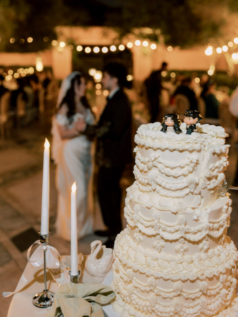 A tall four-tiered white wedding cake with elaborate ruffle and swag buttercream piping, topped with custom Disney animator-style cartoon cake toppers of the bride and groom, displayed beside tall taper candles and a miniature ballet shoe figurine at the One&Only The Palm Dubai wedding reception.